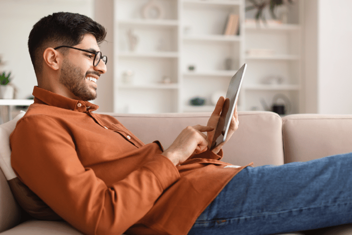 Brown skinned man in an orange shirt and jeans, casually laying on a sofa and looking at an iPad