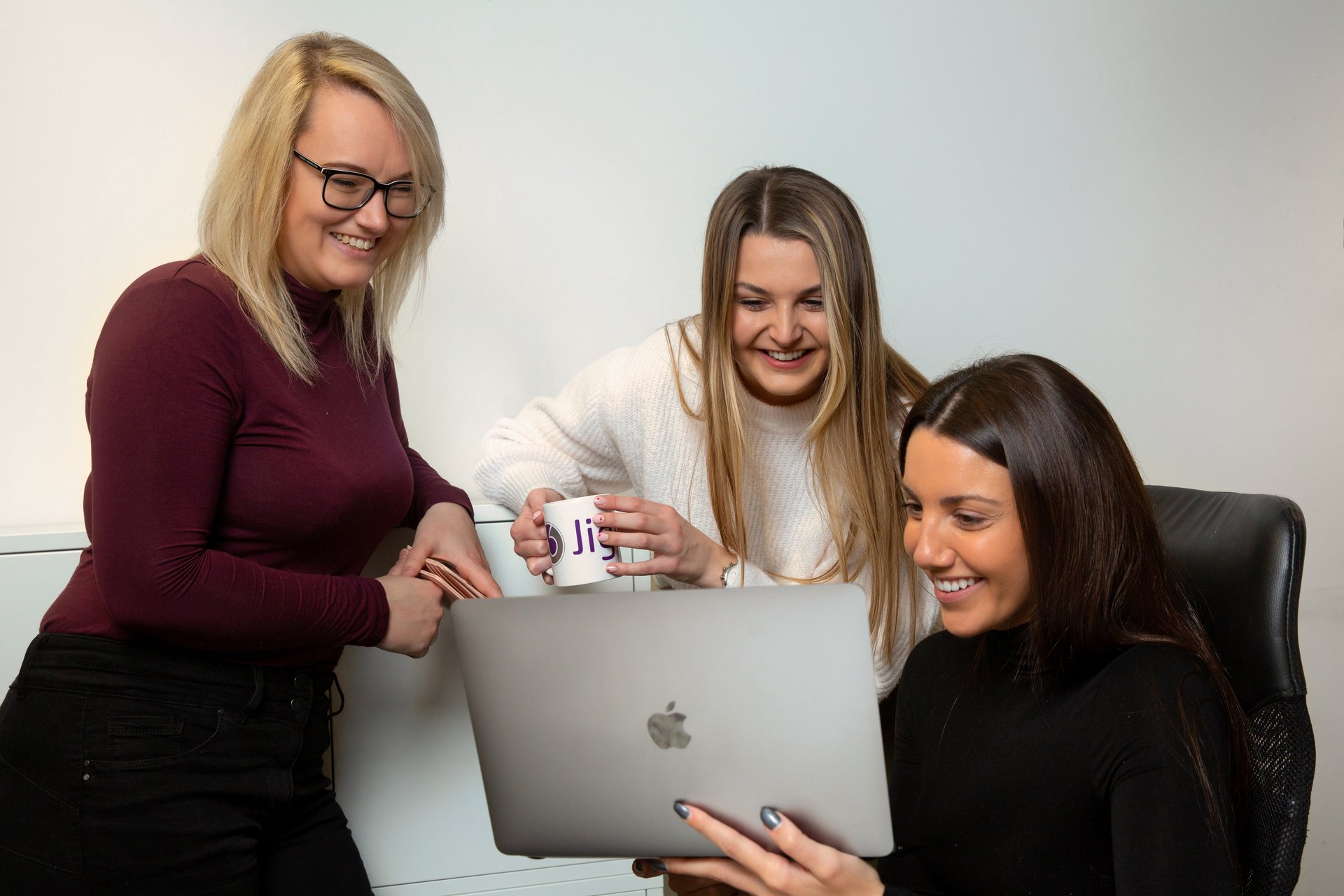 Three women stand around a laptop smiling