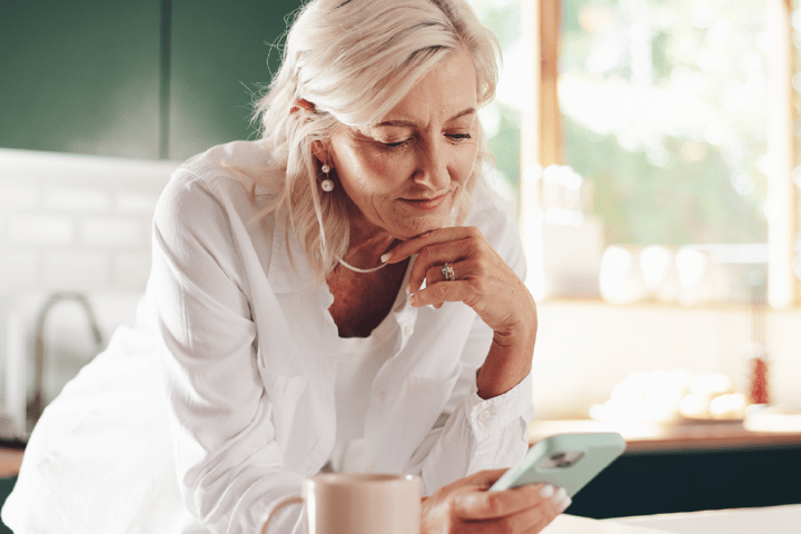 An older lady with wavy blonde hair and tanned skin looking at her phone. She has a mug to the side of her and has her hand placed under her chin. An older lady with wavy blonde hair and tanned skin looking at her phone. She has a mug to the side of her and has her hand placed under her chin.