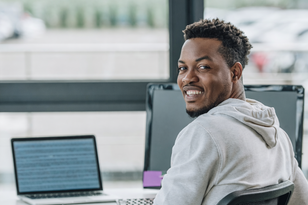 Black-skinned man who has his head turned to face the camera. He is smiling whilst on his laptop Black-skinned man who has his head turned to face the camera. He is smiling whilst on his laptop